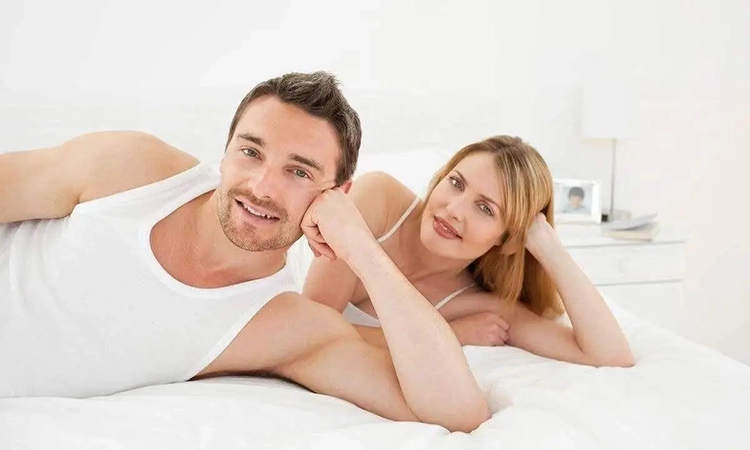 The couple lies on the bed with one hand supporting their head.