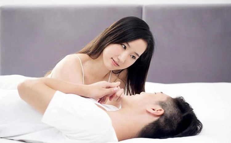 On a bed, a woman is lying on her stomach and looking down at her boyfriend.